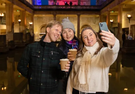 Three visitors taking a selfie in front of the Great Bath, two of them holding hot chocolates.