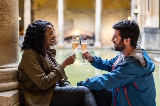 A woman and man sitting by the Great Bath, toasting with glasses